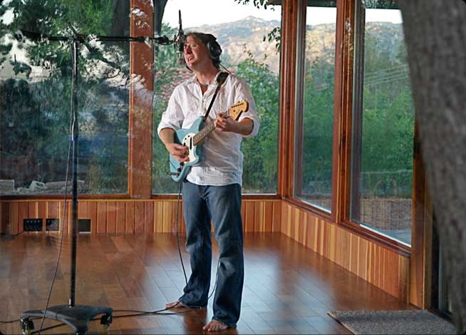 Musician recording in beautiful studio setting, mountains visible through large glass windows in background.